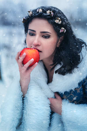 Portrait of young woman dressed in blue coat. She is walking in the winter forest holding a basket with applesの写真素材