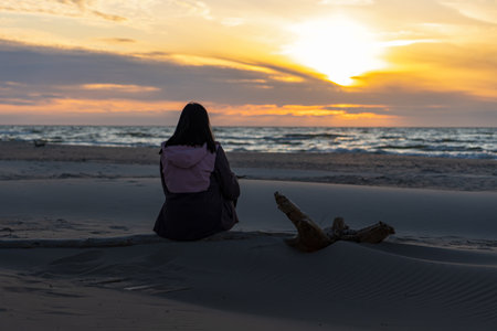 Young woman sits on old driftwood on a sandy beach, watching the sea during a colorful sunset.の写真素材
