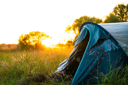 Tent pitched on green grass during sunrise, bathed in warm orange light, with trees in the background creating a cozy outdoor camping atmosphere.の写真素材
