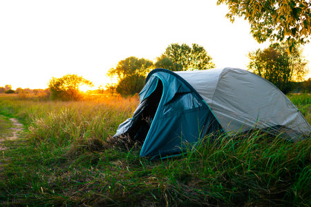 Tent pitched on green grass during sunrise, bathed in warm orange light, with trees in the background creating a cozy outdoor camping atmosphere.の写真素材