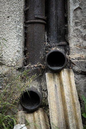 Two vintage metal pipes fixed to a buildings exterior, channeling rainwater from the roof, showing signs of age and weather exposure.の写真素材