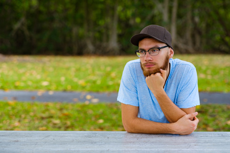 Handsome young man with a beard sitting on a park picnic table.の写真素材