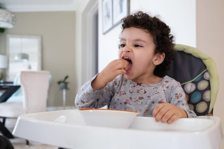Cute two year old baby girl sitting on a high chair feeding herself in a home settingの写真素材