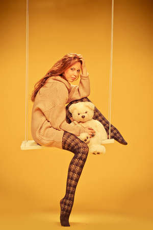 a beautiful young girl sitting on a swing with a bear in his hands in the studioの写真素材
