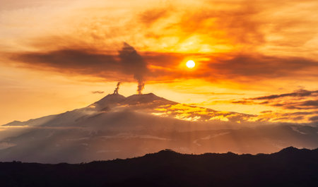 mysterious landscape of great erupting volcano with smoke from craters , clouds of gas and snow on slopes in orange light of sunset. eruption of vulcanの写真素材