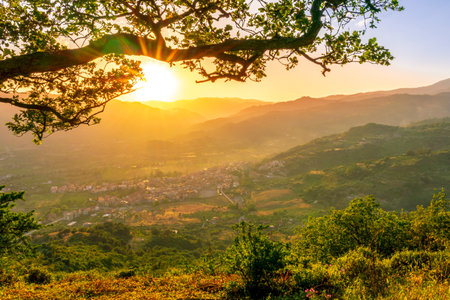 wonderful spring sunset in mountains with amazing view from highland among greeen tree branches to a valley with town, mountains and beautiful sunset on backgroundの写真素材