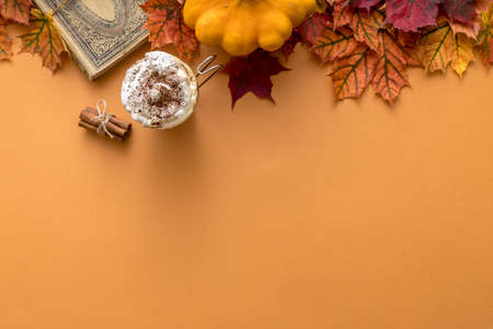 Cozy autumn style flat lay of hot pumpkin spice latte with whipped milk cream, cinamon and colorful dry leaves of october on the light orange background with empty space for logo or designの写真素材