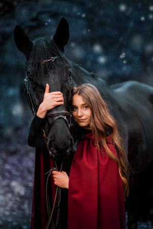 Fabulous portrait of a beautiful young lady with long brown hair, suite with red or burgundy cloak with her black horse in winterの写真素材