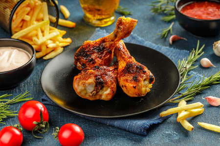 Still life of three grilled chicken legs with crunchy golden brown crust served on a black plate with thighs, vegetables and sauces on a blue textured tableの写真素材