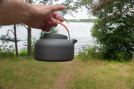 Person with tea kettle stands by lake near trees and grass