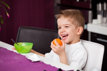 A five year old boy sitting at a kitchen table with an orange in his hand and a bowl in front of himの写真素材