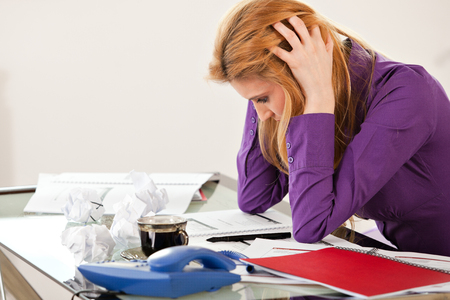 businesswoman holding hands on her head, sitting at desk, with rumpled paper, coffee, red file  and phone, headacheの写真素材