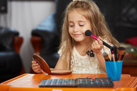 blonde little girl learning the make-up, holding a brush, smiling, looking in the mirrorの写真素材