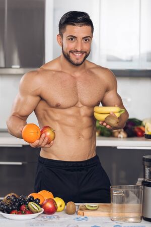 young fit man with fruits, in the kitchen, holding banana, orange, apple, kiwiの写真素材