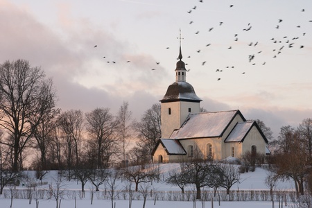 An old Swedish country church surrounded by a winter landscape at dusk with a flock of birds flying by の写真素材