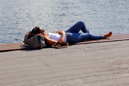 Copenhagen, Denmark - August 24, 2017: An attractive woman wearing blue jeans, white linen has put the shoes next to her when she is laying next to the water in Nyhamn with her head on a bag in the sunshine.のeditorial素材