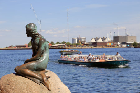 Copenhagen, Denmark - June 27, 2018: The statue of the Little Mermaid, by  Edvard Eriksen, located at Langelinie, with sightseeing boats behind.のeditorial素材