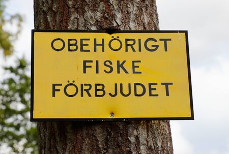 Close-up of a yellow sign with black text in Swedish on tree trunk with information that unauthorized fishing prohibited.の写真素材
