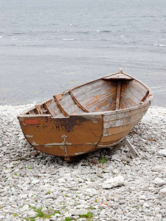 one small open brown wooden boat for fishing seen from the stern resting on rocky shore with the sea in the background.の写真素材
