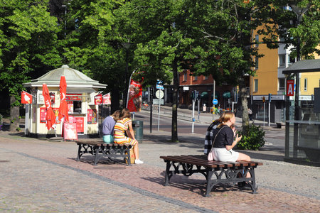 Karlstad, Sweden - June 30, 2021: Four young people are sitting in the sunshine on the benches placed in the square in front of the ice cream kiosk.のeditorial素材