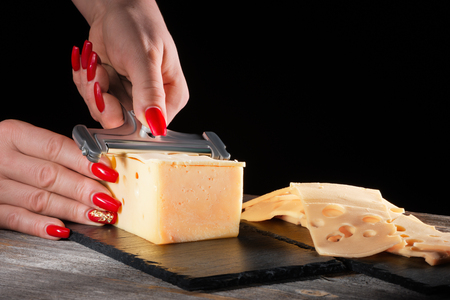 Female hands with beautiful bright manicure cut cheese whetstone by means of a cheese slicer on a black slate plate in style a rusticの写真素材