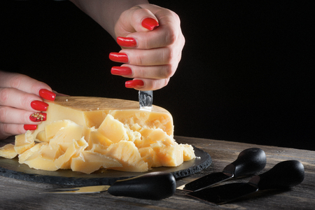 Female hands with bright beautiful manicure break away pieces of hard cheese like parmesan by means of a special knife on a plate from black slate in style a rustic.の写真素材