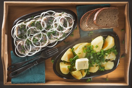 Pieces of a herring with rings of onions and fennel, hot boiled potatoes with green onions and a piece of butter on two ceramic plates of an ovoidny form on a wooden tray from a bambooの写真素材