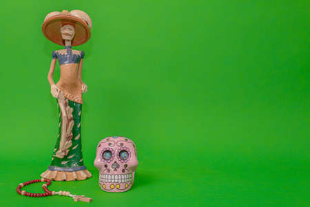 Calavera de la Catrina statuette, Mexican symbol of Day of the Dead, next to a wooden rosary and a pink skull, on a uniform green studio backgroundの写真素材