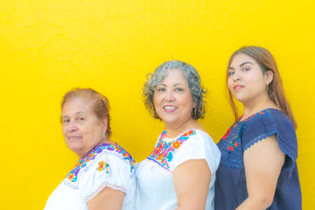 Grandmother, daughter and granddaughter with floral print blouses in a row looking at the camera, three generations of Mexican women on a yellow backgroundの写真素材