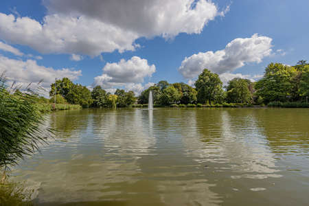 Lake with a fountain with a jet of water surrounded by lush green trees in the city park of Sittard, sunny summer day with a blue sky and white clouds in South Limburg, the Netherlandsの写真素材