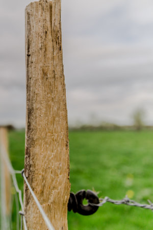 Close-up of a wooden post with wire on a fence bordering a farm field next to a footpath, green grass and gray cloudy sky in the blurred backgroundの写真素材