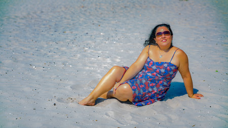 Beautiful latin woman with long black hair in a blue dress with details in red, wearing sunglasses and smiling in the camera, enjoying the sun on a sunny day on the beach in the Netherlandsの写真素材