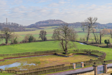 Pond with very little water between a fence of dry bushes, green grass and trees, a country road and the Dutch countryside in the background, blue sky and white clouds in South Limburg, Netherlandsのeditorial素材