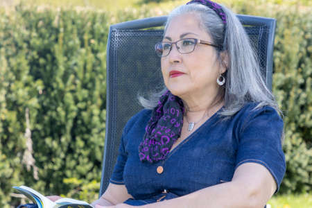 Pensive and reflective mature woman sitting on a garden chair wearing casual clothes, denim dress,...