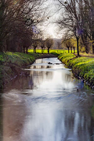 Small river with slight reflection on its waters among green grass and bare trees, green field in the background, sunny autumn day in Sweikhuizen, South Limburg, Netherlands. Long exposure photographyの写真素材