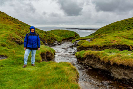 Stream flowing to sea in the Irish countryside, coastal hiking path from Doolin to Cliffs of Moher, hiker woman in blue clothing looking at camera, cloudy rainy day in County Clare, Irelandの写真素材