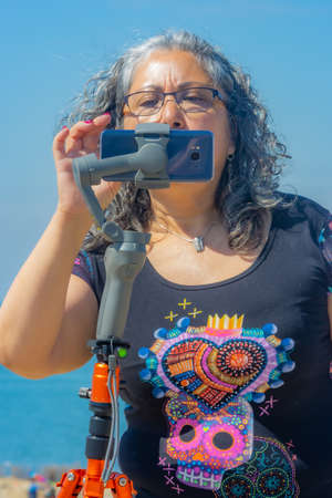 Mature woman with gray hair, glasses filming with her mobile phone gimbal tripod head stabilizer,...