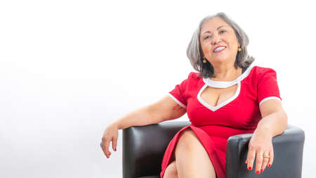 Elegant smiling mature Mexican woman sitting on a bench and looking at the camera, grayish black hair, red dress with white details, white studio background. Successful business woman. Space for textの写真素材