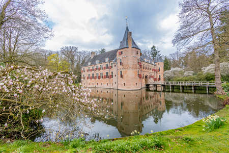 Moat with reflection in the water surrounding Het Oude Loo castle with its bridge between trees and green vegetation, cloudy day with abundant clouds in Paleispark Kroondomein, Apeldoorn, Netherlandsのeditorial素材