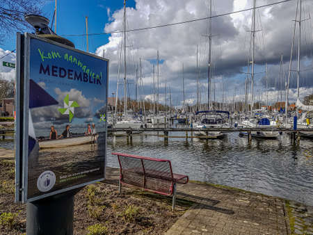 Medemblik, North Holland, Netherlands. April 14, 2021. The city seaharbor, tourist information sign, red metal bench, anchored ships, masts and stormy clouds in the background, calm and cloudy dayのeditorial素材
