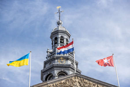 Dutch, Ukrainian and Maastricht flags waving, tower of the town hall building, clock, bells and a weather vane, blue sky with white clouds in the background. Maastricht, South Limburg, Netherlandsの写真素材