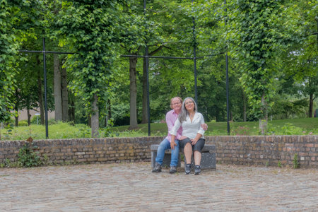 Mature couple sitting on bench embracing, smiling expression, looking at camera, casual clothing, glasses, relaxed day in public park with trees in background in Heerlen, South Limburg, Netherlandsの写真素材