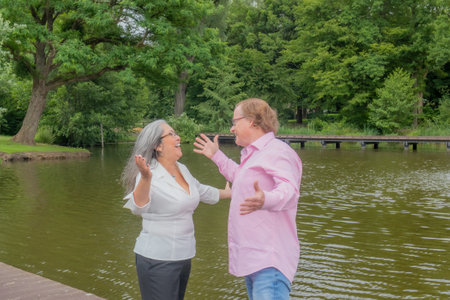 Mature couple with wide smile, arms outstretched to hug each other, casual clothes, glasses, relaxing day in a public park, a pond and trees in the background in Heerlen, South Limburg, Netherlandsの写真素材