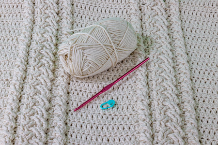 A cotton ball, pink metal hook and green stitch marker on a beige cotton yarn crochet blanket,...