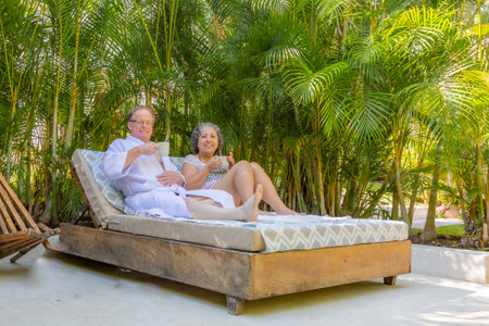 Mature tourist couple sitting on sun lounger against lush green palm trees, smiling and drinking coffee, looking at camera, beachwear, short gray hair, enjoying relaxed sunny summer day at spaの写真素材