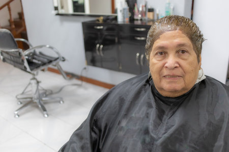 Old woman with dye on her head sitting on a chair in beauty salon with a blurred background, short hair, expression with a slight smile, front view. Old age fashion and beauty concept. Space for textの写真素材