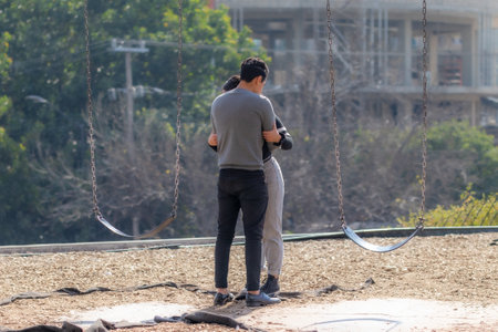 Guadalajara, Jalisco Mexico. December 28, 2022. Romantic scene of young woman and young man embracing between two swings, buildings in blurred background, backs to camera, sunny day in the cityのeditorial素材