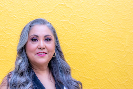 Frontal portrait of senior adult woman looking at camera over yellow background, grey-black hair, warm color makeup, lips with pink lipstick, formal wear, black and white blouseの写真素材