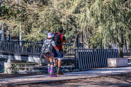 Zapopan, Jalisco Mexico. January 1, 2023. Couple a woman and a man on roller skates on a sidewalk with a bridge and a tree with green foliage in background, sunny day for two to enjoy outdoor sportsのeditorial素材