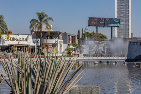 Guadalajara, Jalisco Mexico. January 8, 2023. Cityscape seen through Glorieta Minerva fountain, palm trees, trees, billboards and buildings in blurred background, sunny day with a blue skyのeditorial素材